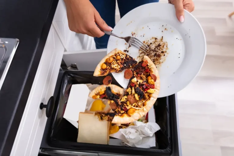 Hands using a fork to scrape uneaten food off of a plate into a kitchen trash bin.